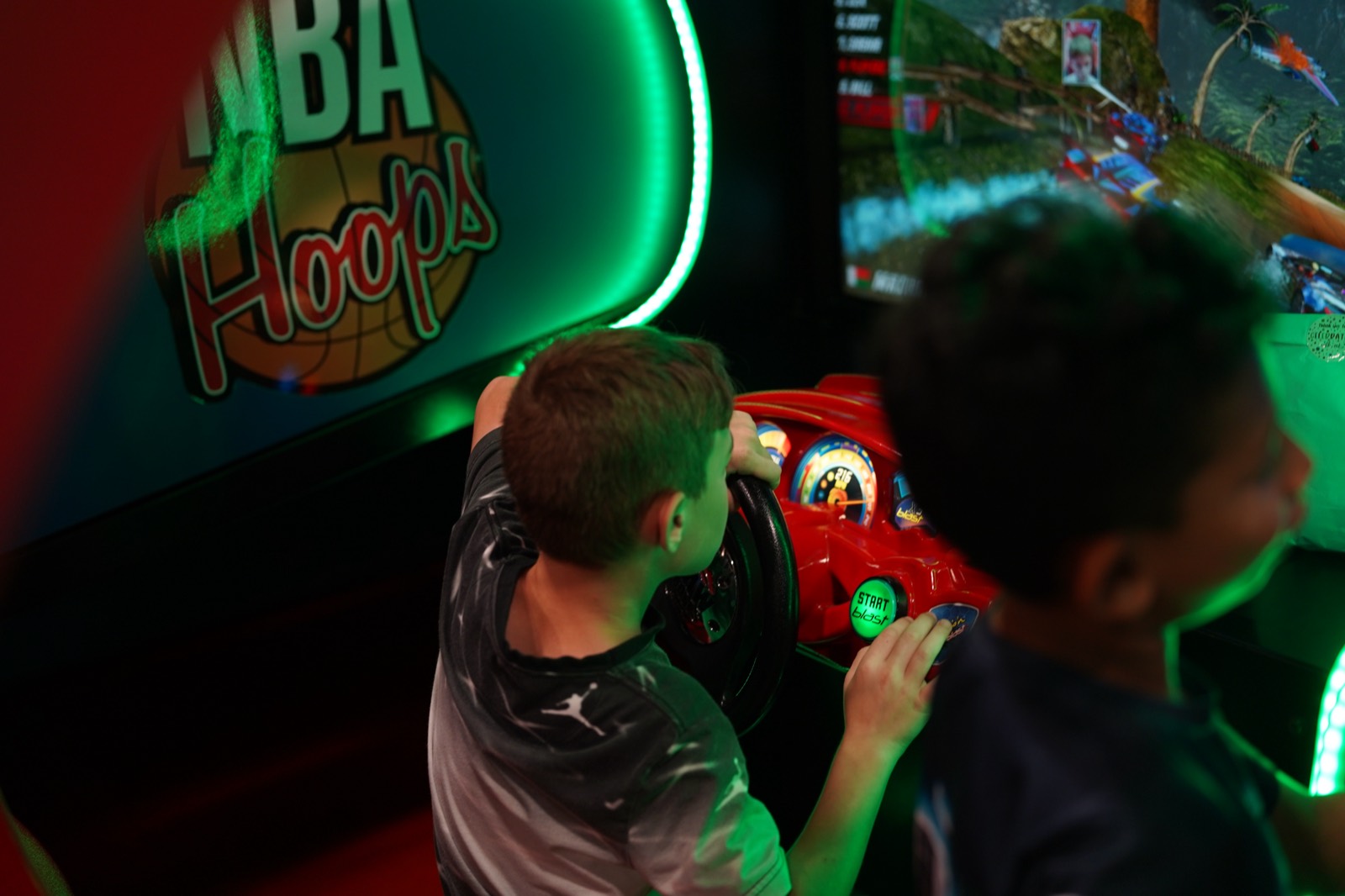 Two kids playing a sit-down driving racing cabinet beside NBA Hoops at the SkyTag arcade
